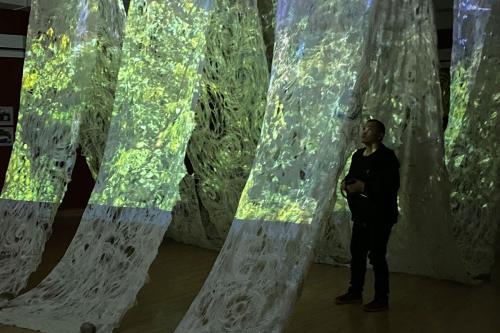 Textile installation hung from the ceiling. A visitor in all black walks under it, looking up.