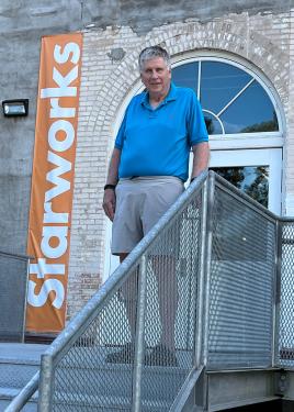 A man in a blue polo shirt with gray graying hair stands at the top of an exterior stairway, at the entrance to a brick building with an orange Starworks banner by the door.