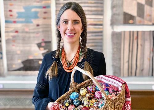 A woman in a dark blue blouse and two long brunette braids holds up a woven wicker basket full of intricately designed and dyed Easter eggs.
