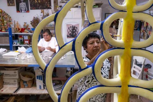 Two women working in an artist workshop. In the foreground, the older woman paints the bare “branches” of a Tree of Life sculpture. In the background, seen through the branches, the other woman molds clay.