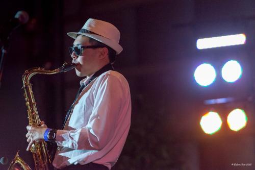 An Asian man wearing fedora and sunglasses plays saxophone under bright stage lights.