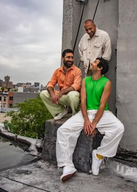Three Arab men pose on a rooftop, smiling at each other, with a low city skyline and gray skies in the background.