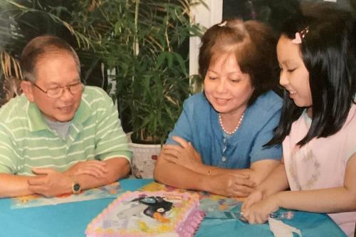A young girl in a pink heart T-shirt smiles down at the pink decorated cake on the table in front of her. An older man and woman sit next to her, also smiling at the cake.