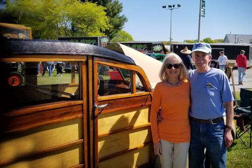 A woman and man pose arm in arm next to an old wood-paneled car.