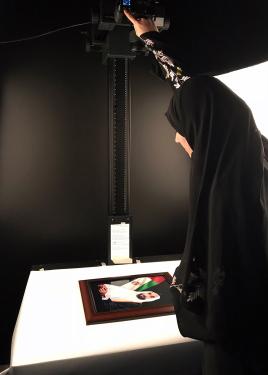 A person in black hijab, facing away, adjusts an overhead camera to shoot a color photograph on a white table.