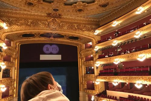 Golden interior of an opera hall, with several tiers of box seats, still lit before a show begins. A child in the foreground faces away, looking up at the tiers.