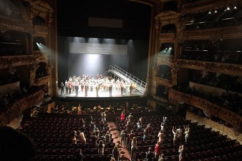 Scattered members of an audience give a standing ovation for performers taking bows on stage in an opera hall.