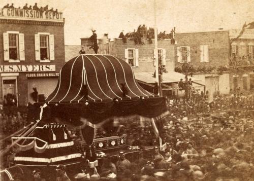 Crowds of people around what seems like a parade float, containing a coffin. There are even people on the roofs of the buildings watching.