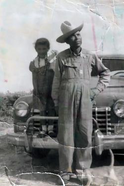 A young girl stands on the fender of an old truck alongside her father in overalls and cowboy hat. The black-and-white photo has been crinkled and stained.