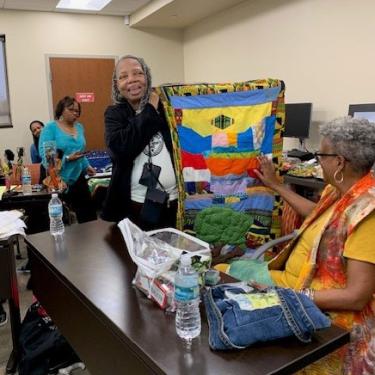 A woman holds up a small quilt with brightly colored shapes. Another woman grazes her hand over it.