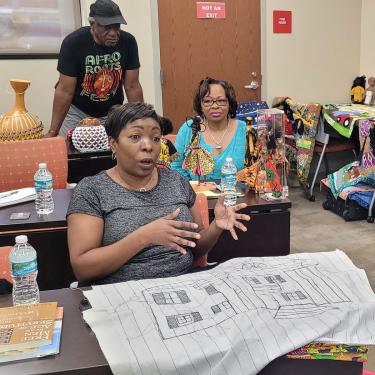A woman, seated at a table in a workshop, gesticulates over a fabric pattern of a building.