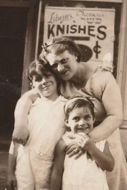 A woman leans down, posing with her arms around two smiling children, in front of a storefront sign that reads Litwin’s Famous Knishes, 5 cents. Black-and-white photo.