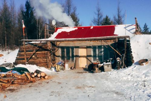 One-story cabin with red roof and walls of stacked timbers to protect from snow drifts. Snow covers the ground and part of the roof. Smoke rises from a chimney, over a small satellite dish.