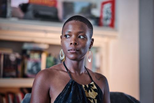 A woman wearing a  black halter top and silver earrings looks off to the side, a bookshelf blurred behind her.
