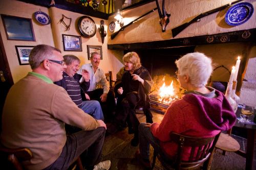 Five people sit in a circle in front of a lit fireplace. One woman is speaking and gesticulating while the others laugh.