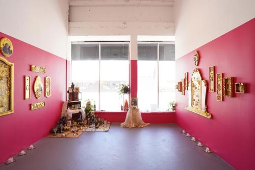 Gallery walls painted pink, lit by large windows. In one corner, an altar with frame photos and potted plants. On the walls, gold framed artwork.