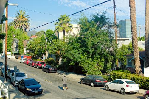 Photo from a second-story porch overlooking a street lined with parked cars, palm trees, and other green foliage. A man crosses the street.