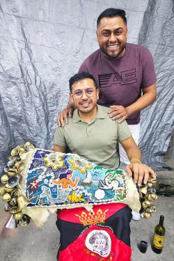 One man sits holding a decorated drum, while another stands behind him with his hands on his shoulders. Both smile at the camera.