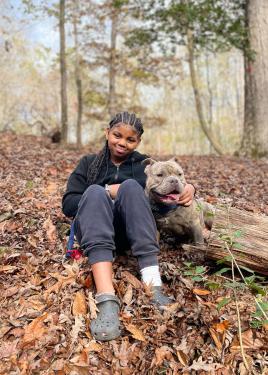 A girl wearing black hoodie, sweatpants, and Crocs sits among fallen brown leaves on the ground, one arm around a gray-brown pitbull.