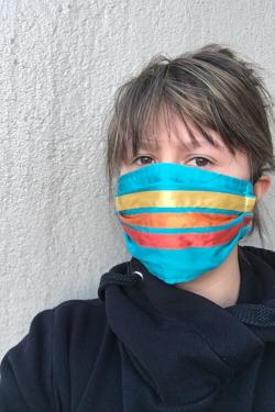 Portrait of a woman in a black sweatshirt and a turquoise face mask with three parallel ribbons sewn across it: yellow, orange, red.