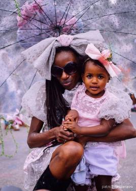 A Black woman crouches down, embracing her daughter. They both wear pale pink frilly dresses and large bows in their hair.