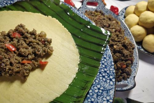 Ingredients for pastelles in the making: a small heap of ground meat and vegetables in the center of a circle of dough, on top of a green banana leaf. In the  background, more ground meat and balls of dough.