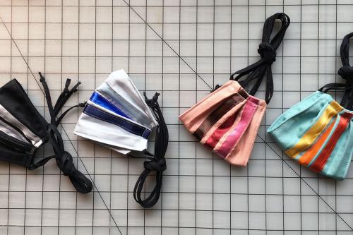 Four face masks on a gridded workstation. They have black straps and stripes of ribbons in contrasting colors.