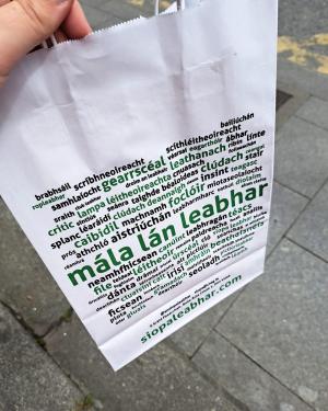 A white paper shopping bag, printed with several words and phrases in the Irish language.