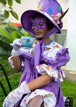 A Black man dressed in purple frilly dress, oversized purple bonnet, and lacey white gloves, seated, daintily tips from an large tea cup. 