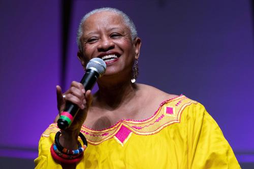 A woman with short gray hair and a bright yellow blouse speaks into a microphone on stage, smiling.