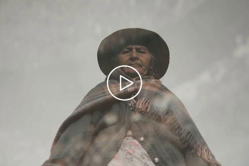 A film still of an elderly woman wearing a wide brimmed, black hat, wrapped in a woolen poncho. The image looks up at her from below.  On top of the image is a play symbol.