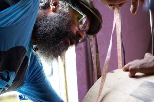 A man with beard, glasses, and pageboy hat leans over, measuring a wooden drum.