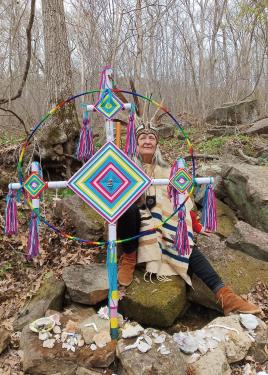 A woman wearing a woven poncho holds up a colorful God’s eye marker among bare trees in the woods.