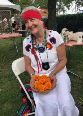 A woman wearing white embroidered dress, turquoise necklaces, and a red headband sits in a folding chair at an outdoor festival, holding a bowl of orange marigolds.