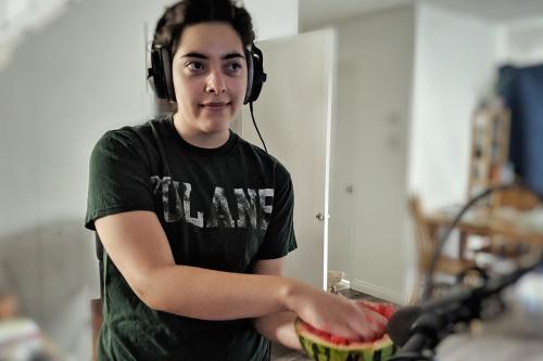A woman wearing large headphones smirks as she squishes the flesh of half a watermelon with one hand, her eyes fixed on a screen out of frame. A black microphone is pointed at the watermelon.