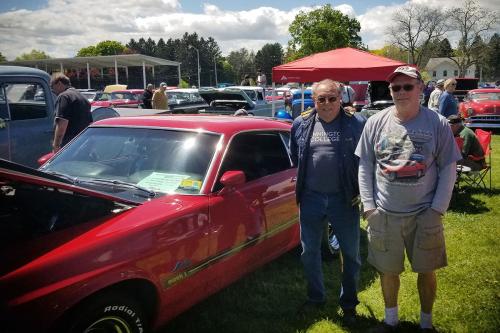 Two men pose in front of a classic red car with the hood popped.