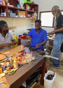 An elder man and two young men work in a workshop, at a table set with measuring tools, scissors, containers of glue, and scraps of leather.