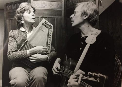 A woman playing autoharp and a man playing acoustic guitar face each other as they sing together. Black-and-white photo.
