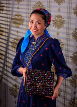 A woman poses wearing a dark blue embroidered dress, blue  headscarf, and holding up a shoulder bag.