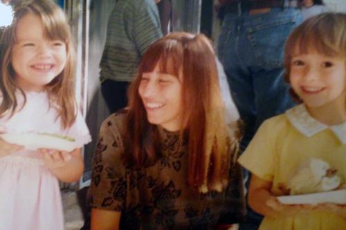 A woman kneels down, eyes closed and smiling, with two toddler girls on either side of her holding paper plates of cake.
