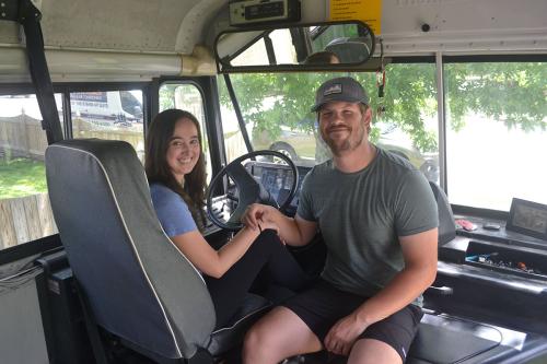 A woman, sitting in the driver's seat of a school bus, bumps fist with her partner sitting next to her.