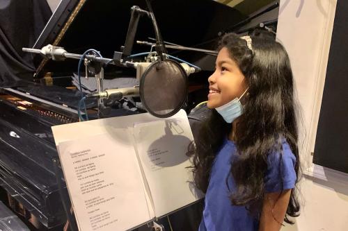 A young girl stands at a microphone and music stand containing printed lyrics, in front of a grand piano.