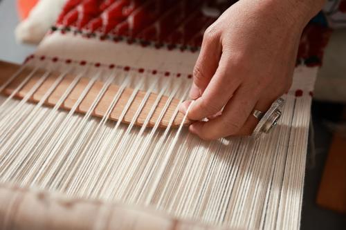 Close-up on a hand, wearing a large silver ring, leading a wooden shuttle through alternating white threads on a loom.