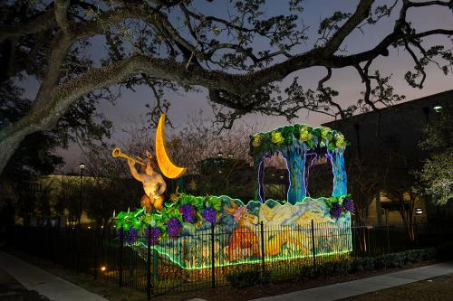 A yard decorated with a forest scene: animals, foliage, and a mythical creature sounding a trumpet under a yellow crescent moon. At night, the edges of the sculpture are lit up in green.