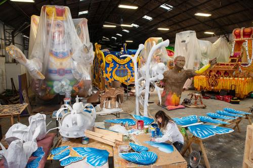 In a warehouse, a woman paints prop pieces on the ground, with several other float pieces behind her.