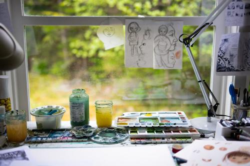 Detail of the artist's desk, with a window looking out toward a blurred forest. The desk is set with watercolor palettes, water containers, jars of brushes, and a lamp. A few illustrated outlines are taped up in the window.