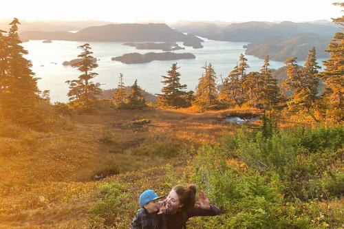 A young woman and young boy pose, each with one hand raised, in front of a meadow and body of water at golden hour.