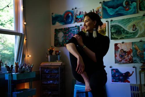 A woman poses in her art studio, perched on a stool, wearing all black and woven earrings. Behind her are art supplies and watercolor illustrations on the wall.