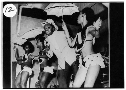 A man in white jacket and women in frilly bikini costumes holding small umbrellas dance on stage.