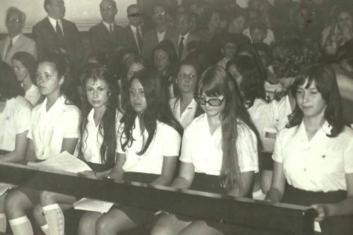 Young women students in uniform sit side by side, with men in suits standing behind them. Black-and-white photo.